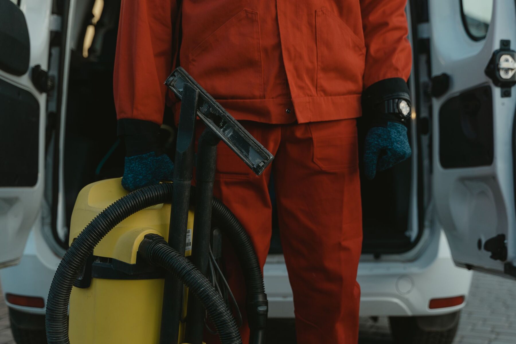 Technician in red coveralls and work gloves holding a wet vacuum and wand at the back of an open service van